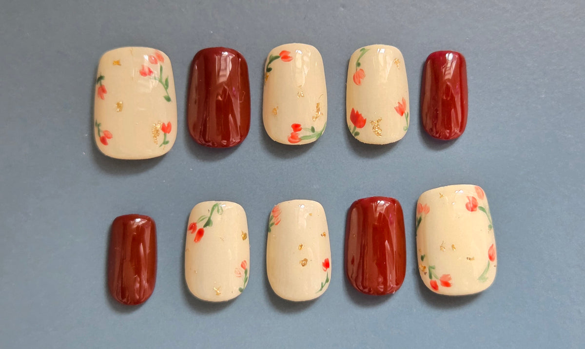 Ruby Meadow hand-painted red floral press-on nails on a cream base with gold foil accents
